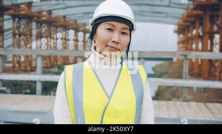 portrait of female builder standing outside construction area wearing safety helmet. People and technical profession concept. Stock Photo