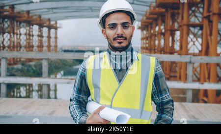 portrait of Arab construction specialist in helmet standing in building site holding plan and looking at camera. People and industry concept. Stock Photo