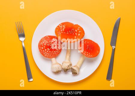 Amanita muscaria fly agaric poisonous mushrooms on plate with fork and knife on yellow background top view. Consumption of hallucinogenic mushroom con Stock Photo