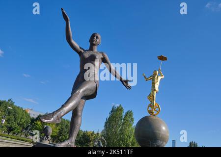 A bronze sculpture of a young woman ballet dancer. A gold sculpture of a boy riding a unicycle is in the background.  In the plaza in front of the Cir Stock Photo