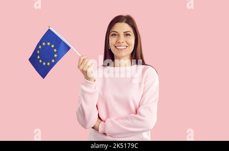 Happy smiling female European Union citizen waving small EU flag on pink background. Stock Photo