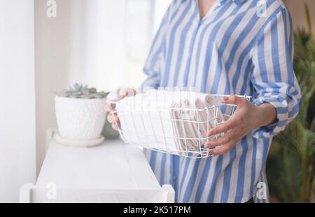 Happy young woman in bathrobe is placing the folded bed sheets in white steel wire basket on shelf Stock Photo