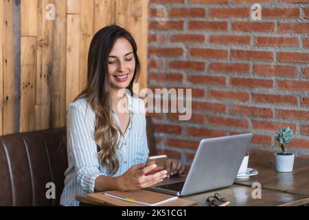 Side view of young happy woman in smart casual clothes browsing on smartphone while working on netbook siting at table and working on remote project i Stock Photo