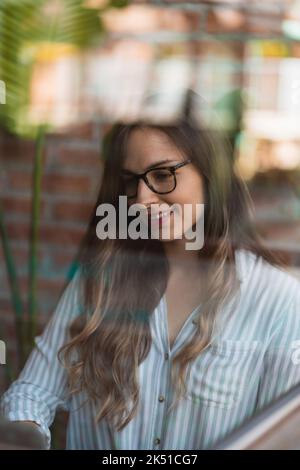 Smart young female manager in striped blouse and eyeglasses with long hair opening laptop while starting to work on business project behind glass in w Stock Photo