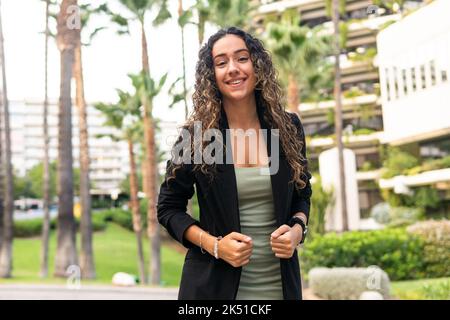 From below self assured young Hispanic businesswoman in stylish black jacket with curly hair looking at camera on blurred background of city street Stock Photo