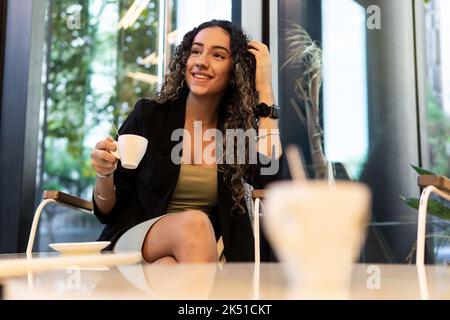From below young Hispanic female in stylish dress touching curly long hair and looking away with smile while sitting in armchair and enjoying coffee i Stock Photo