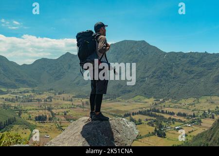 Back view of unrecognizable young male traveler in casual clothes and cap standing with backpack on rocky boulder and admiring scenic mountainous land Stock Photo