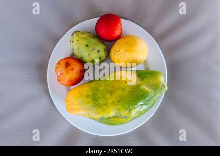 Top view of delicious ripe whole exotic papaya and cactus fruit with mango and plums served on white ceramic plate and placed on bed Stock Photo