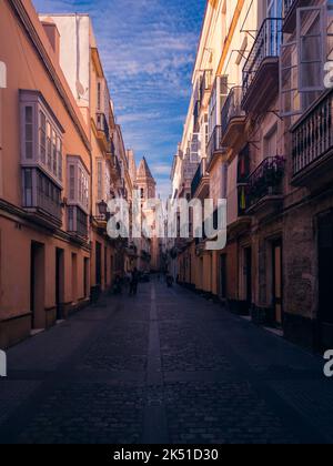 Dark narrow stone paved street with low rise buildings with balconies and cathedral at distance under blue sky Stock Photo