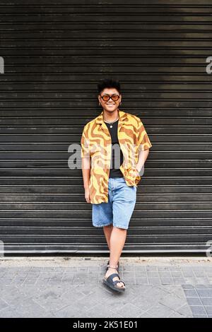 Smiling Filipino male in trendy shirt and sunglasses standing at grunge metal wall with hand on pocket and looking at camera with smile Stock Photo