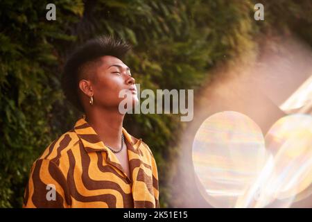 Dreamy young Filipino non binary man wearing cool shirt with earring standing with eyes closed in green garden Stock Photo