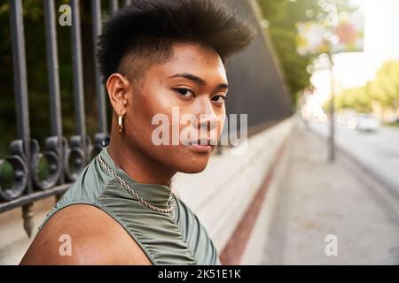 Side view of Asian transgender woman with short hair looking at camera while standing on blurred background of road and park in city Stock Photo