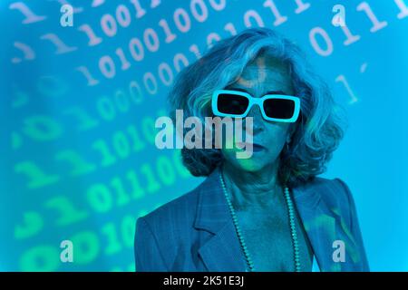 Confident elderly woman in smart casual clothes and sunglasses looking at camera while standing under under turquoise projection of binary code Stock Photo
