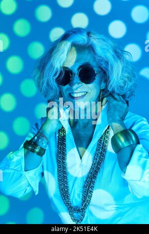 Happy elderly woman with stylish accessories smiling and adjusting collar of white shirt under blue neon light and projection of circles Stock Photo