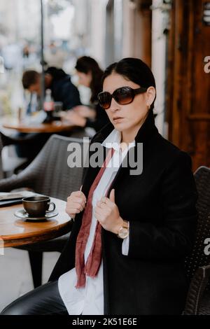 Serious adult Hispanic female with dark hair in white shirt coat and sunglasses sitting near round table of street cafe and looking at camera Stock Photo