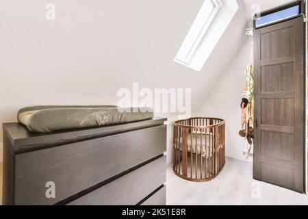 White crib placed at wall near window in light spacious children bedroom with wooden chest of drawers at home Stock Photo