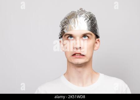 Portrait of anxious young man wearing tin foil hat looking up in panic. Conspiracy theories and paranoya concept. Studio shot on gray background Stock Photo