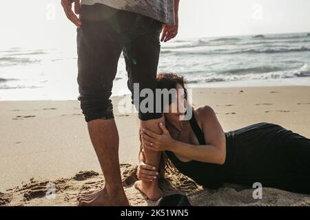 woman in dress lying on wet sand and hugging leg of man near ocean,stock image Stock Photo