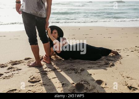 woman in dress and hat lying on wet sand and hugging leg of man near ocean,stock image Stock Photo