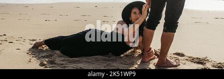 woman in dress and hat lying on wet sand and hugging leg of man near ocean, banner,stock image Stock Photo