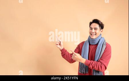 Positive Asian man in red sweater and scarf pointing fingers at ad empty space isolated on beige color background. Cheerful fashionable 20s guy aside pointing gesture - Half body length studio shot. Stock Photo