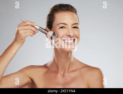 Face, makeup and brush with a mature woman applying blusher to her cheek in studio on a gray background. Beauty, skin and cosmetics with an attractive Stock Photo