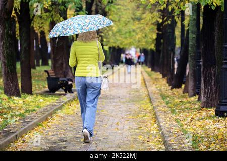 Rain in city, slim woman with umbrella wearing jeans and yellow jacket walking down the street. Rainy weather in autumn park Stock Photo