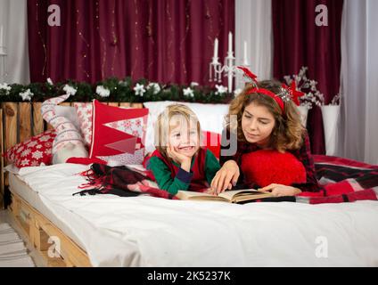 Christmas Eve with family. The older sister and younger brother are lying in pajamas on the bed and reading a book. friendship concept, spending holid Stock Photo