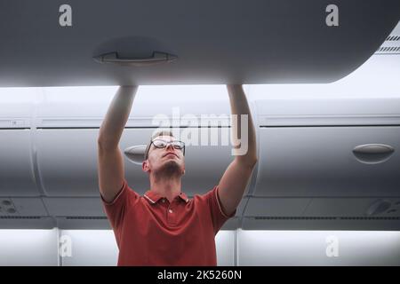 Man travel by airplane. Passenger putting hand baggage in lockers above seats of plane. Stock Photo