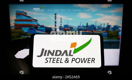 Person holding cellphone with logo of company Jindal Steel and Power Limited (JSPL) on screen in front of webpage. Focus on phone display. Stock Photo