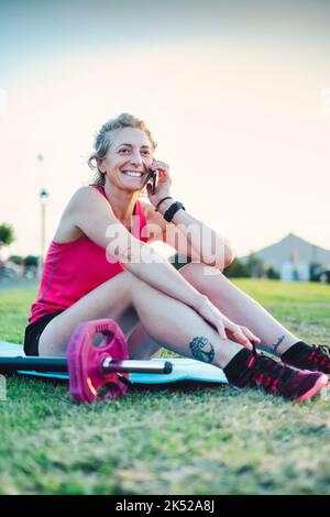 Real young mature caucasian woman in a sport wear talking with her cellphone in the park on a sunny afternoon. Stock Photo