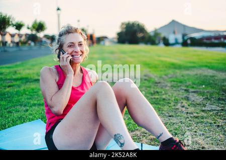 Real young mature caucasian woman in a sport wear talking with her cellphone in the park on a sunny afternoon. Stock Photo