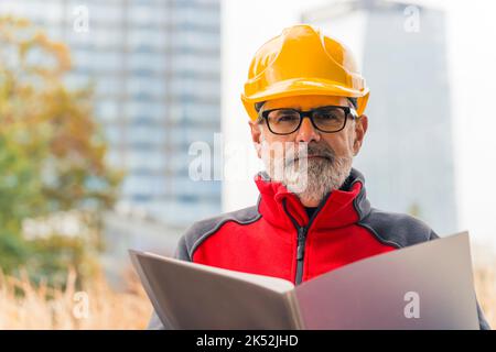 Building industry concept. Adult experienced caucasian male engineer in yellow hardhat looking at camera and holding folder with building plans. Outdoor closeup portrait. High quality photo Stock Photo