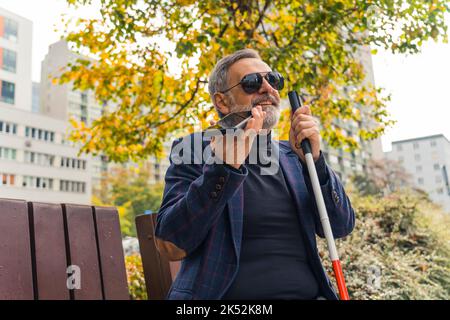 Sight challenged bearded caucasian man in formal clothing smiling, sitting on bench at park, holding walking crane with one hand, and holding smartphone with the other. Live with disability, Outdoor shot. High quality photo Stock Photo
