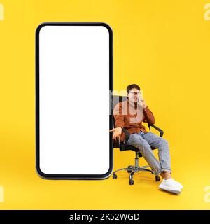 Man Talking On Cellphone Near Large Smartphone Sitting, Yellow Background Stock Photo