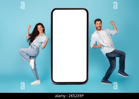 Happy excited young international guy and woman dance, rejoice to win near big smartphone with blank screen Stock Photo