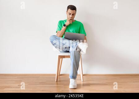 Pensive arab man using laptop and thinking, browsing internet while sitting on chair against white wall at home Stock Photo