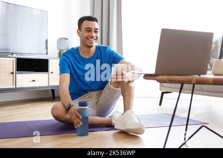 Online workout. Positive arab man exercising at home, looking at laptop, using smartphone, having fitness class online Stock Photo