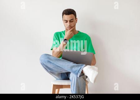 Thoughtful arab guy using laptop while sitting on chair against white wall, working or studying remotely on portable pc Stock Photo