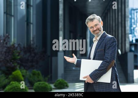 Mature gray-haired teacher invites pupils and students to study, senior male university lecturer looks at camera and smiles invitation gesture with hand, senior lecturer near university building. Stock Photo