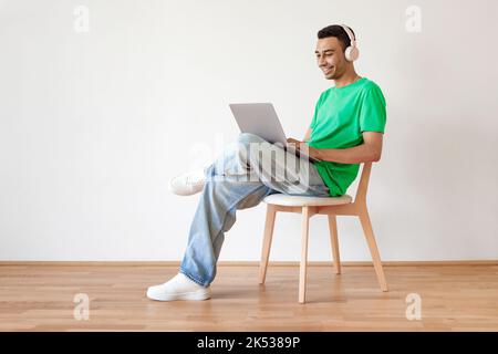 Young arab man using pc, working online on laptop and wearing wireless headphones, sitting on chair over white wall Stock Photo