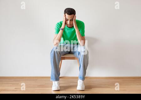Pensive young arab man thinking, touching head with both hands, sitting on chair over light wall, copy space Stock Photo