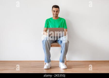 Cheerful arab male freelancer using laptop computer, sitting on chair over white wall and typing on keyboard, copy space Stock Photo