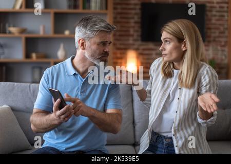 Angry mature caucasian husband points at smartphone and scolds to wife on sofa in living room interior Stock Photo