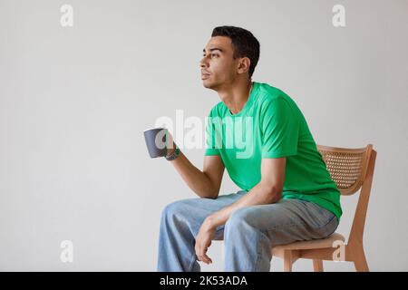 Thoughtful arab man drinking coffee and looking away, sitting on chair over while wall, free space Stock Photo