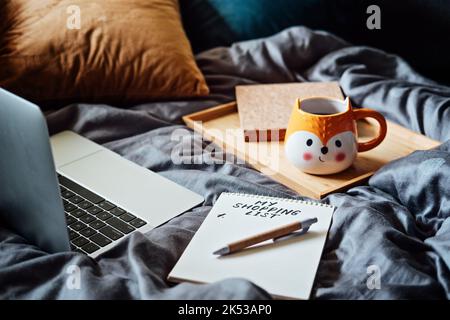Shopping List in note book near laptop and tea coffee cup at bad in home. Shopping Preparation Consumer Buying Stock Photo
