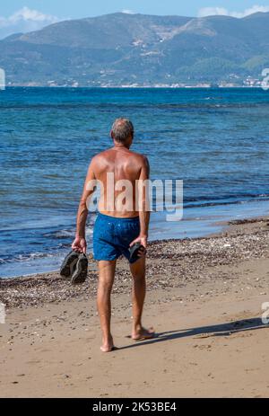 older man walking along a beach wearing blue shorts, middle-aged man walking along a sandy beach on holiday in greece, man walking alone on a beach. Stock Photo