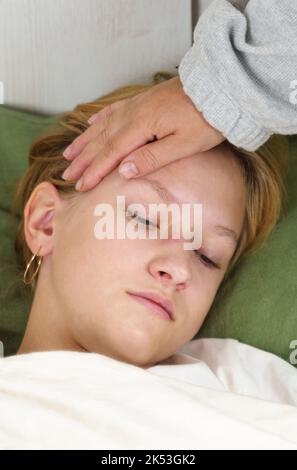 Mom touches her hand to the forehead of a sick daughter lying on the bed. Checks temperature. Vertical frame. The concept of health and medicine. Stock Photo