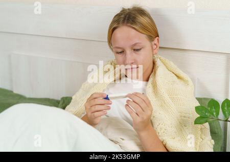 Sick girl sits on the bed and looks at the thermometer. Medicine and health concept. Stock Photo