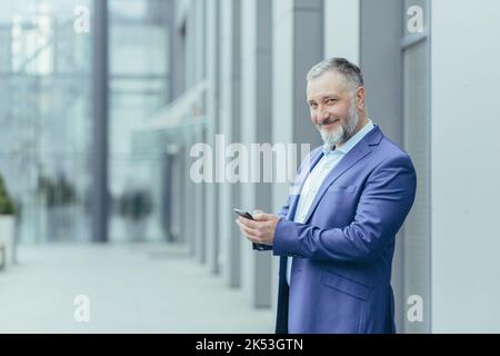 Senior handsome gray-haired man stands in a suit against the background of a modern building, uses the phone, checks calls, mail, messages. He looks at the camera, smiles. Stock Photo